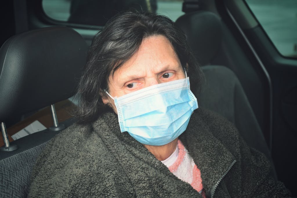 Senior woman wearing a face mask sitting inside a car, looking outside with concern.